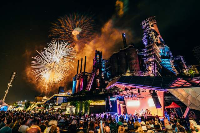 A concert stage at night with a crowd of people in front and fireworks bursting above