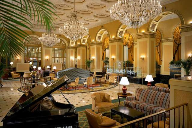 Main lobby of an old, luxurious hotel with seating, elegant chandeliers hanging above, and a grand piano