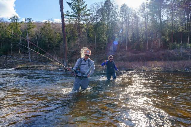 A man and woman in fishing gear wading through a stream holding long fishing poles