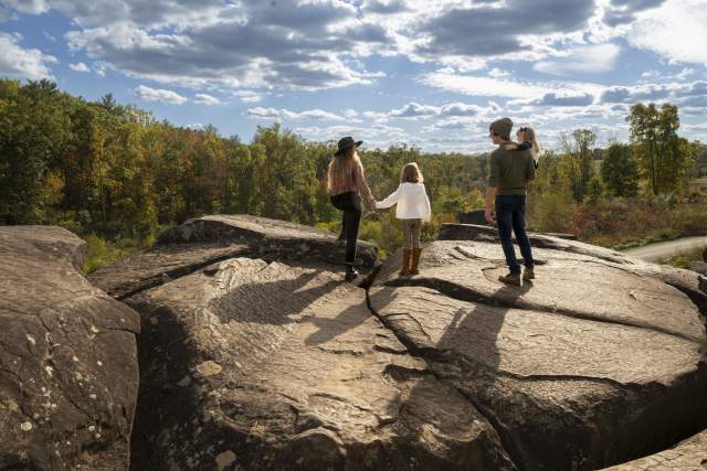 A family standing on a large rock overlooking trees. The woman is holding a young girls hand, while the man is carrying another small child.