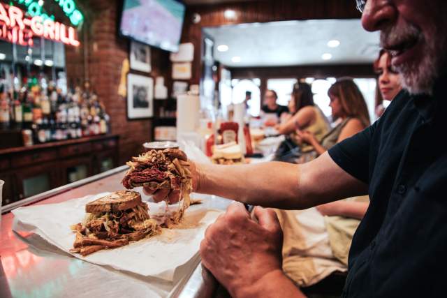 Man sitting at a bar counter holding up a half of a sandwich that has meat, cheese, and french fries piled up