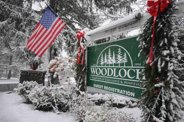 Entrance sign that reads "Woodloch Guest Registration" with pine trees framing either side, an American flag hanging off of it, and snow covers some of the sign and the ground.