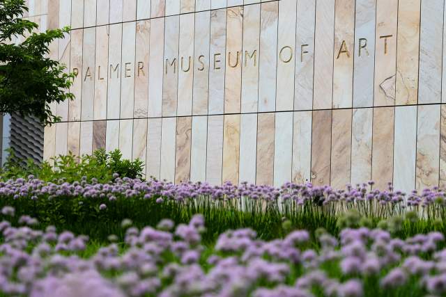 Exterior of a stone museum building that reads Palmer Museum of Art with purple flowers in front
