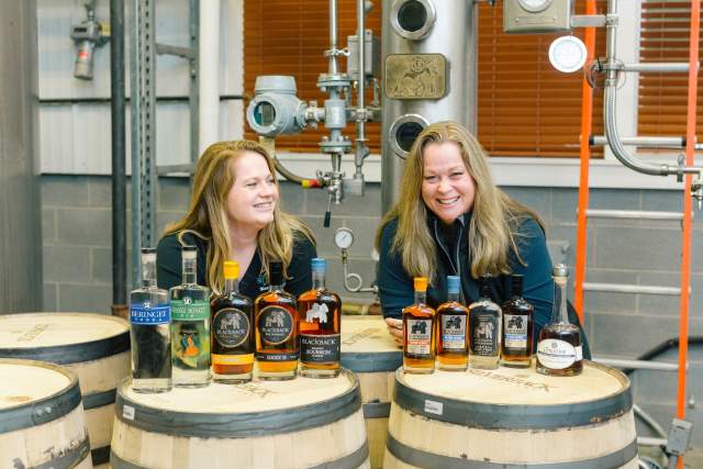 Two women leaning on giant wooden barrels posing behind bottles of gin and bourbon