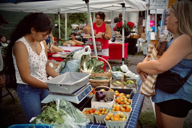 A woman purchasing produce at a farmers' market table while another woman is helping her pay