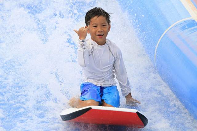 Kid riding a boogey board on a water ride as he makes the hang loose sign with his hands