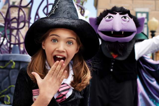 Little girl in a witches hat with her hand over her mouth smiling. A purple puppet is in the background.