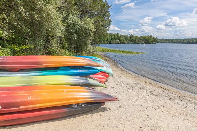A pile of kayaks on top of one another on the sandy shore of a lake