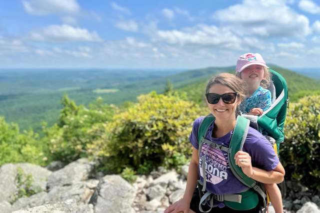 A woman with a young girl strapped to her back on top of a scenic overlook on a sunny day.