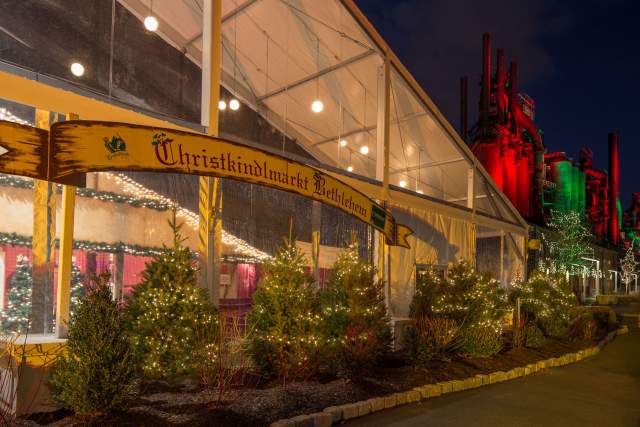 Exterior of a giant see through tent that reads "Christkindlmarket Bethlehem" with Christmas trees aligned beneath the sign.