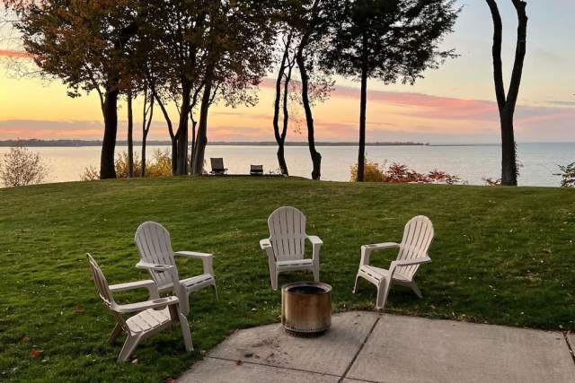 Adirondack chairs surrounding a small firepit on a patio while a sun is setting on a lake in the background