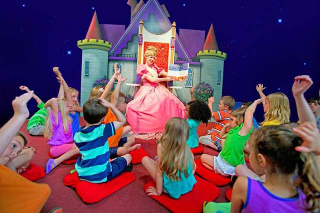A princess reading a book to a group of kids sitting down and raising their hands