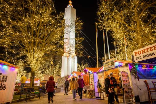 People walking outside at night at the Easton Winter Village in the Lehigh Valley with holiday lights on trees and street vendor tents.