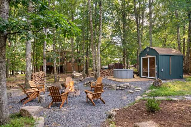 A backyard of a cabin in the woods with Adirondack chairs around a firepit, string lights, and a hot tub