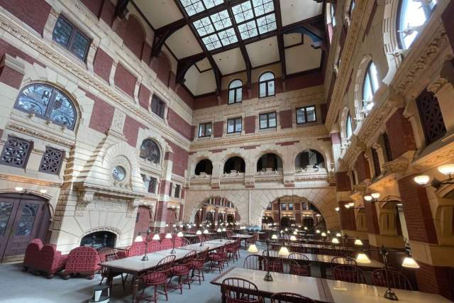 Spacious room with high ceiling, brick walls, arched windows, and a large skylight. Furnished with long tables, red chairs, and lamps.