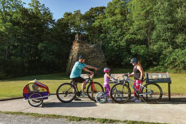 A family of three standing with their bikes around a historical plaque in front of an old furnace
