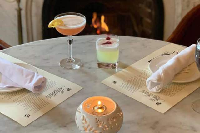 A set table with two placemats, two cocktails, and a candle in front of a fireplace.
