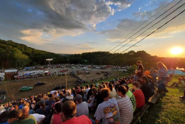 Groups of people sitting on a hill watching cars race and crash into one another in a dirt field