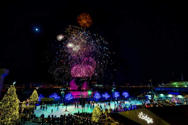 Fireworks bursting in the dark sky above people ice skating on a rink that is surrounding by lit up Christmas trees