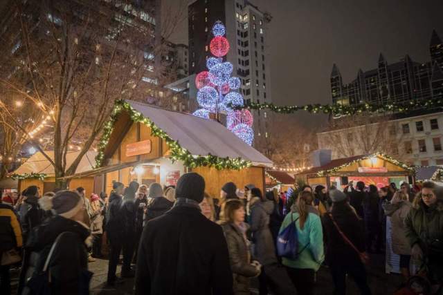 People bundled up at the People's Gas Holiday Market in Pittsburgh at night with holiday lights on the vendor cottages.