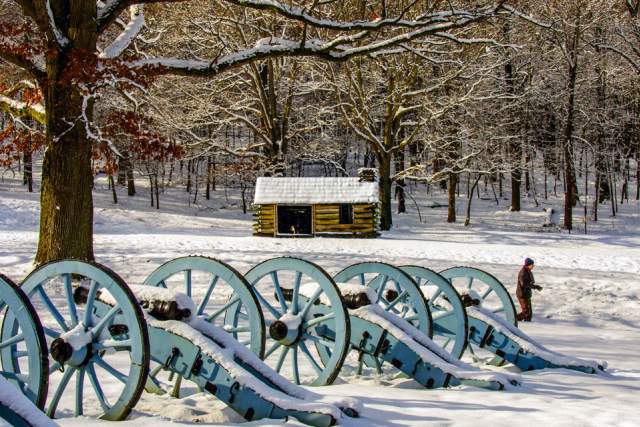 Three old canons in a tow on snow covered ground with a small log cabin covered in snow in the background