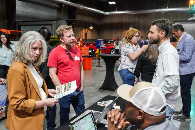 Man and woman standing talking to a vendor at a table. In the background are more people and classic cars