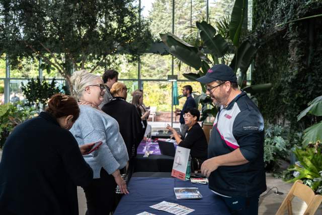 People standing at a conference table talking in a room with a wall of windows and lots of plants. The table has brochures and tabletop stands.