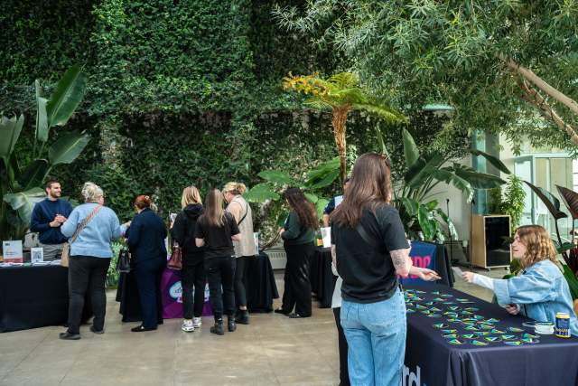 Open and airy greenhouse with tables set up in it and people standing at those tables interacting with the people seated there.