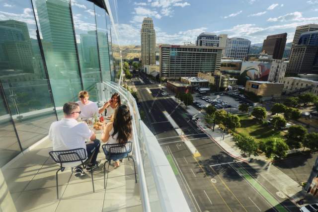 4 people sitting at a rooftop patio table with a glass building on the left and a glass railing on the right with the buildings and scenery of downtown Salt Lake