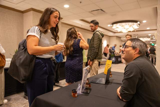 Woman standing at a vendor table talking to the man seated at the table. Busy crowd in the background