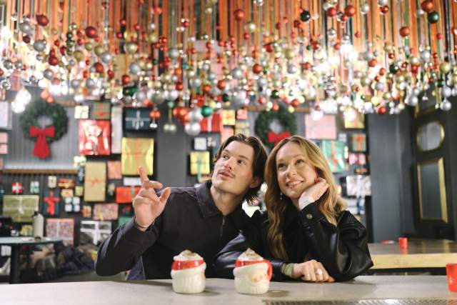 A couple looking whimsical with santa mugs and holiday ornaments hanging from the ceiling