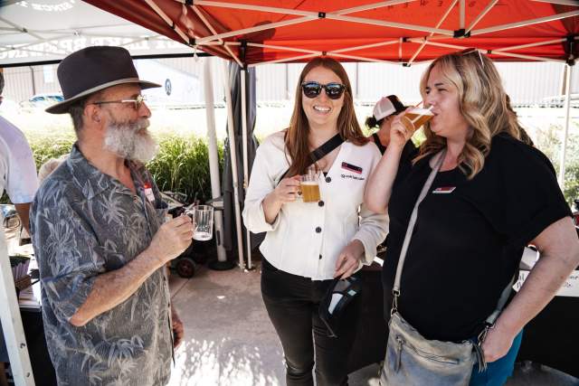Three people standing under a red outdoor shade smiling and drinking beer from clear plastic cups