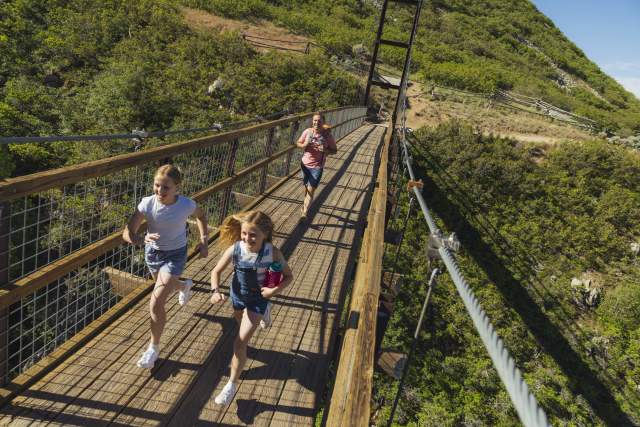 Father and two girls running across a walking path suspension bridge with green mountainside in the background