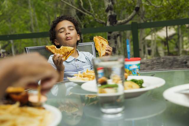 Kid with a very satisfied look on his face holding a sandwich. He is on a patio dining area with trees in the background