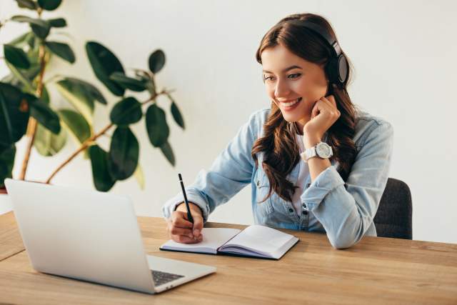A woman wearing headphones and taking notes while watching a webinar on a laptop.