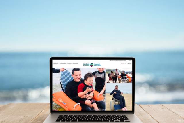 A laptop open to Northern Ireland's Inclusive Beaches page sits on a doc in front of a body of water