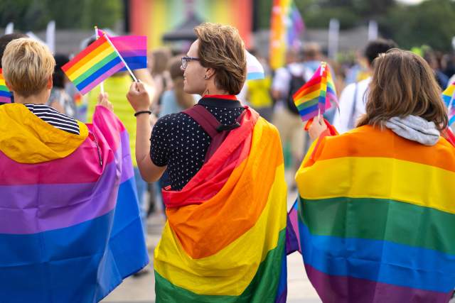 A trio of people wrapped in Pride flags stand at a parade.