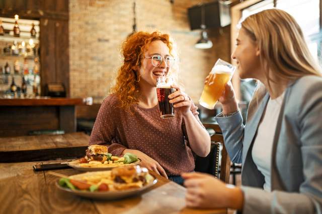 Two women sit at a bar sharing a beer and plates of food