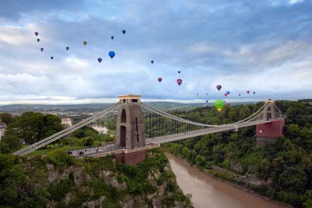 Hot air balloons hover over a bridge spanning a river.