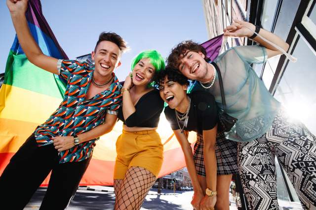 Young LGBTQ+ people celebrating pride together. Group of young queer people smiling cheerfully while holding the rainbow pride flag. Four friends celebrating together at a gay pride parade.