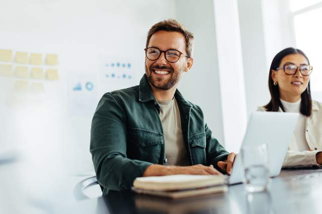 Happy man wearing glasses and a blue shirt listening to a discussion in an office boardroom.