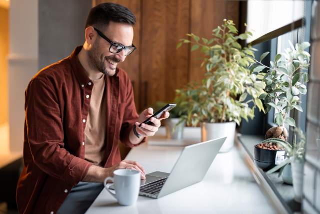 A man in a brown shirt and glasses looks at an email on his phone while sitting at a desk.