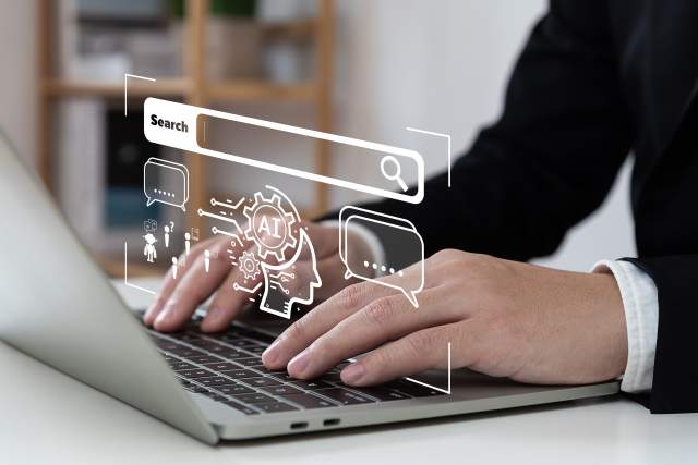 A man's hands type on a laptop while a graphic of an AI chat bot floats, superimposed onto the image.