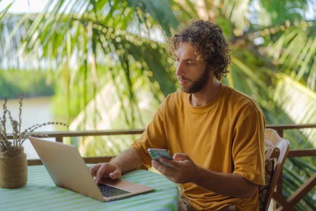 A man sits on a patio outdoors, surrounded by palm trees, while doing work.