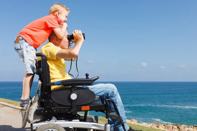 A young boy stands on the back of his father's wheelchair as he looks out with binoculars.