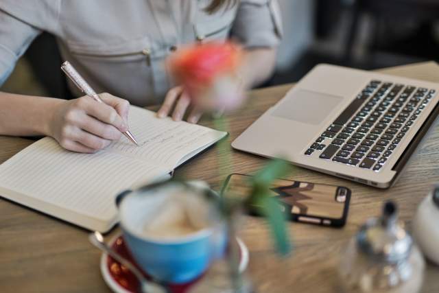 Person Writing On A Notebook Beside Macbook