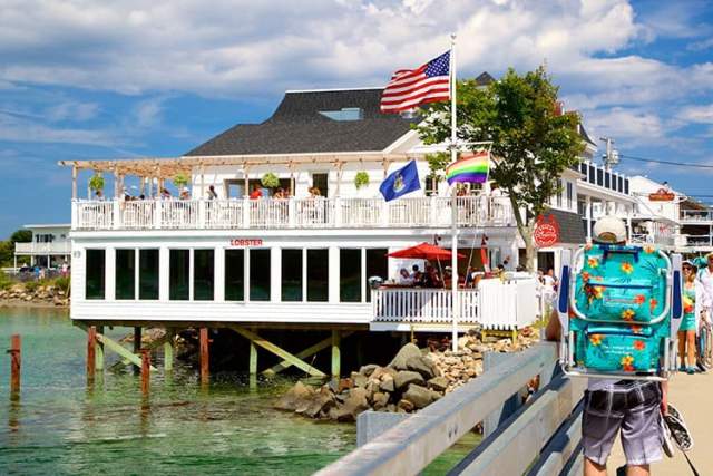 Visitors walk along a waterfront pier past a restaurant overlooking the water in York County, with outdoor seating, flags, and clear coastal views.