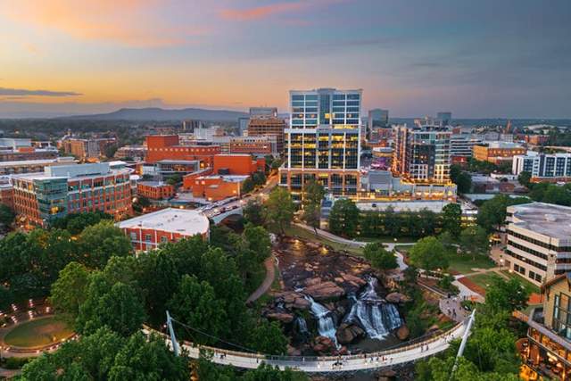Downtown Greenville, South Carolina at sunset, featuring a waterfall, curved pedestrian bridge, trees, and illuminated city buildings.