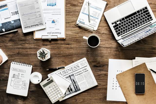 An assortment of items, like a newspaper, calculator, cup of coffee, laptop and notebook, on top of a desk.