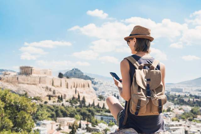 A person sitting on some rocks overlooking some ruins looks down at their phone.